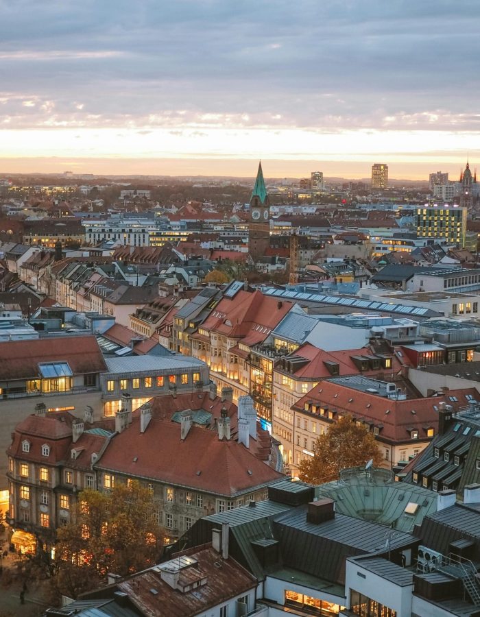 Vue aérienne d'une ville au coucher du soleil avec un mélange de bâtiments modernes et historiques, des toits rouges, des lampadaires lumineux et une église avec un clocher vert au centre.