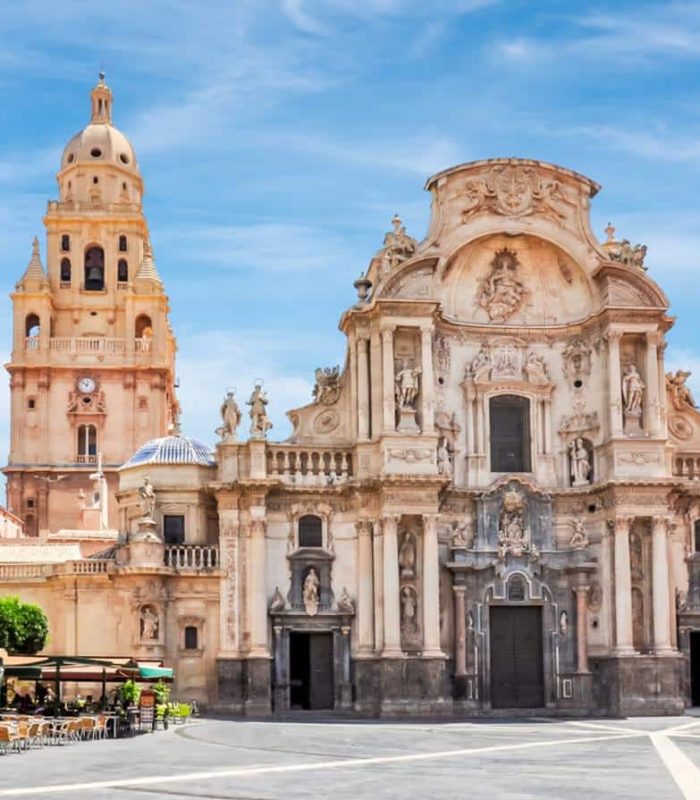 Façade de style baroque de la cathédrale de Murcie, avec ses détails ornementaux, ses statues et son clocher central, sur fond de ciel bleu, sur une vaste place avec, à gauche, des places de café en plein air.