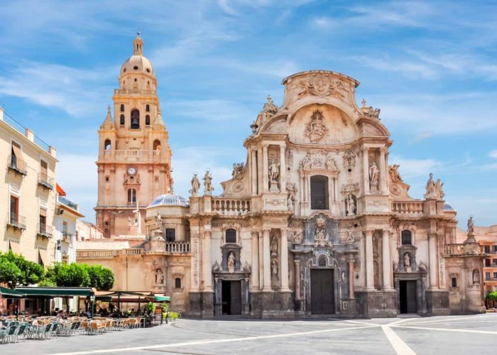 Une grande cathédrale baroque ornée de sculptures et de statues se dresse sur une place ensoleillée, avec un haut clocher en arrière-plan et des sièges de café en plein air sur la gauche.