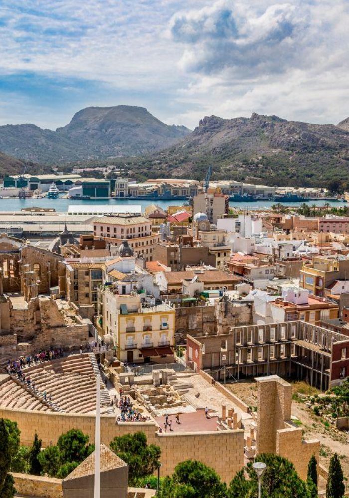 Vue panoramique de Carthagène, Espagne, montrant l'ancien théâtre romain au premier plan, des bâtiments colorés, un port et les montagnes environnantes sous un ciel partiellement nuageux.