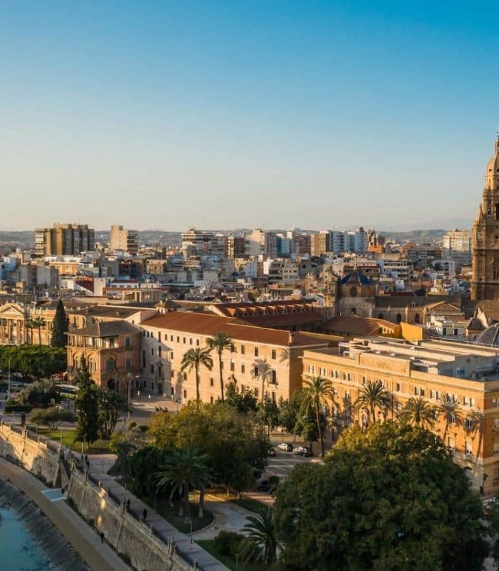 Vue aérienne d'une ville avec une cathédrale proéminente, des bâtiments historiques, des appartements modernes et une rivière avec un pont piétonnier sous un ciel bleu clair.