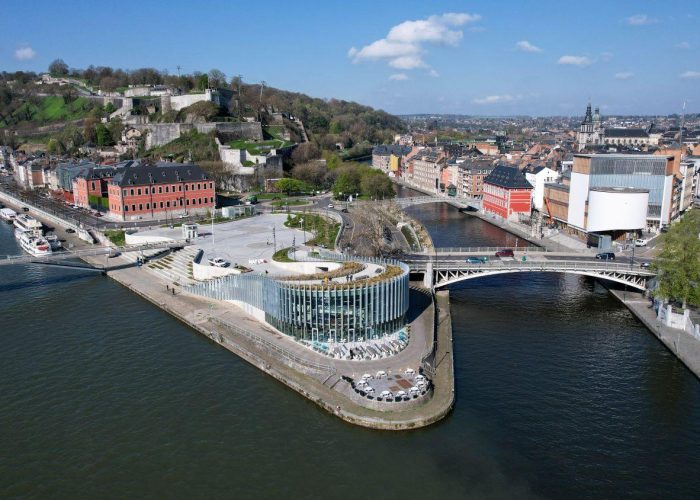 Vue aérienne d'un bâtiment moderne en verre sur une péninsule fluviale, entouré de ponts et d'une architecture urbaine historique, avec une colline verdoyante et un fort en arrière-plan, sous un ciel bleu avec des nuages épars.