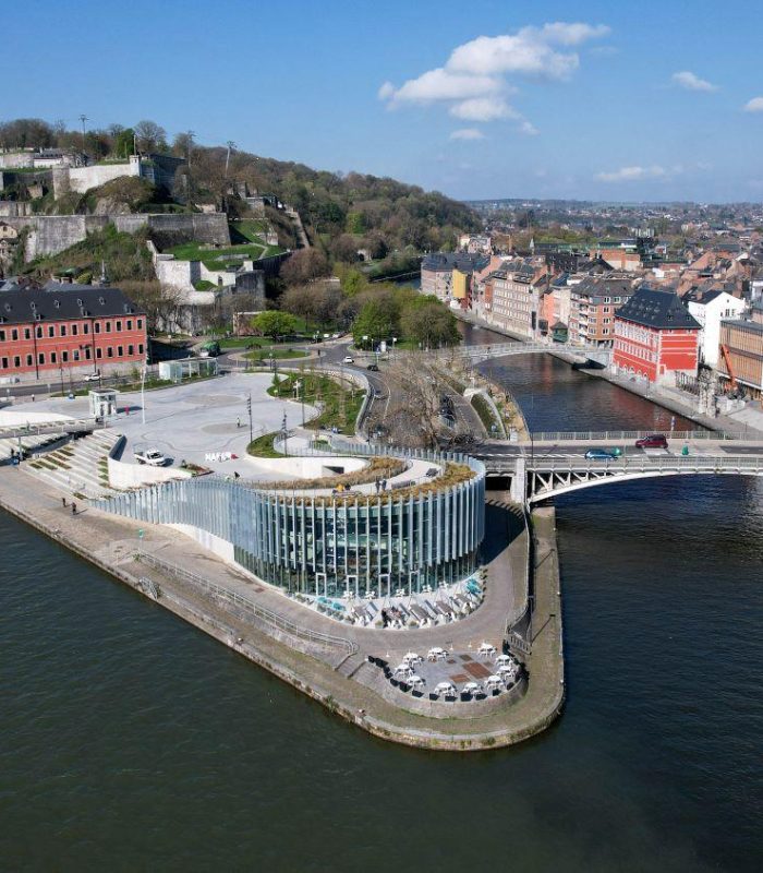 Vue aérienne d'un bâtiment moderne en verre sur une péninsule fluviale, entouré de ponts et d'une architecture urbaine historique, avec une colline verdoyante et un fort en arrière-plan, sous un ciel bleu avec des nuages épars.