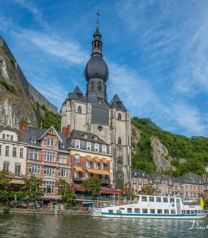 Vue pittoresque de Dinant, en Belgique, avec des bâtiments colorés, l'église collégiale Notre-Dame et son dôme en forme d'oignon, et un bateau blanc amarré sur la Meuse, sous une colline rocheuse.