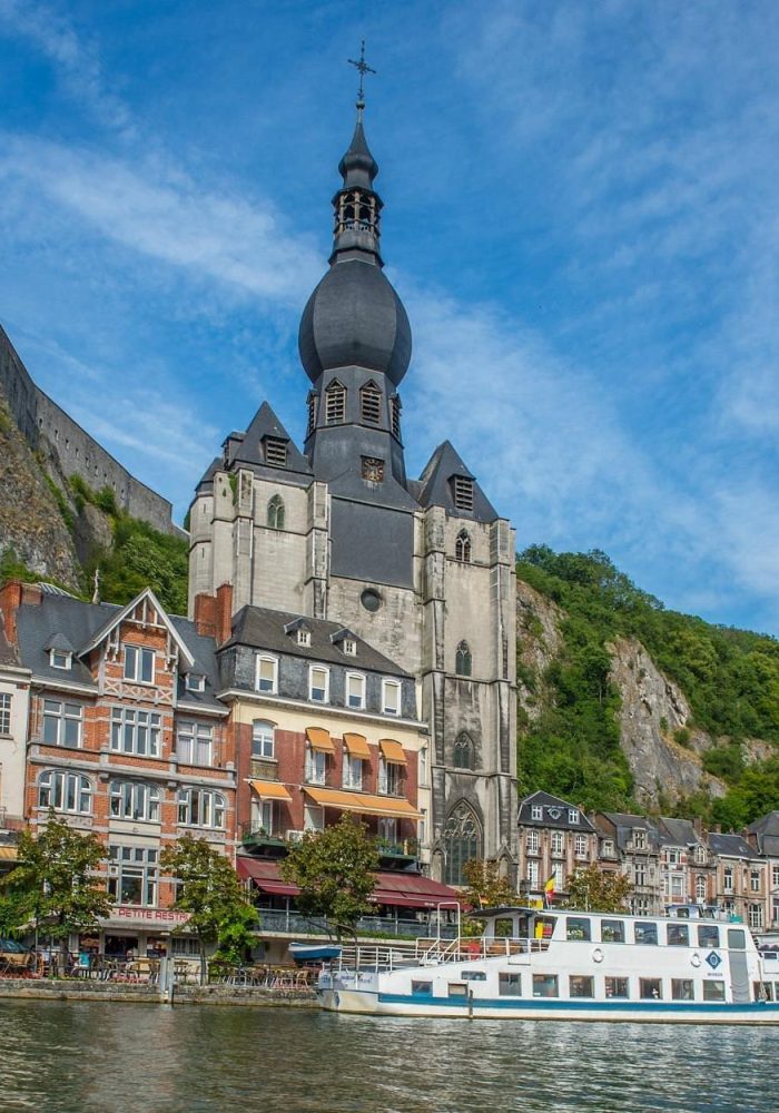 Vue pittoresque de Dinant, en Belgique, avec des bâtiments colorés, l'église collégiale Notre-Dame et son dôme en forme d'oignon, et un bateau blanc amarré sur la Meuse, sous une colline rocheuse.