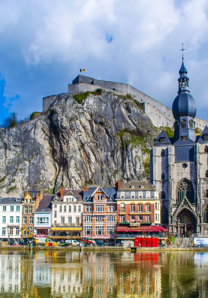 Colourful buildings line the waterfront in Dinant, Belgium, with the ornate collegiate church of Notre-Dame and a fortress perched on a rocky cliff, all reflected in the calm river under partly cloudy skies.