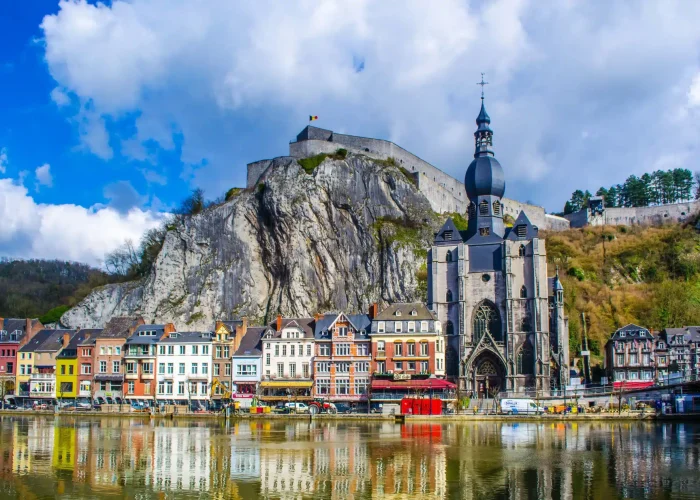 Des bâtiments colorés bordent le front de mer à Dinant, en Belgique, avec la collégiale ornée de Notre-Dame et une forteresse perchée sur une falaise rocheuse, le tout se reflétant dans la rivière calme sous un ciel partiellement nuageux.