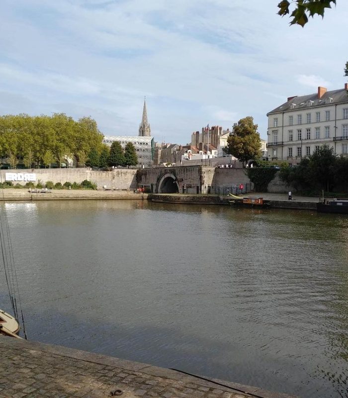 A calm river with a small sailing boat moored at the edge of the stone, trees and historic buildings line the opposite bank, with a church spire visible in the background under a partly cloudy sky.