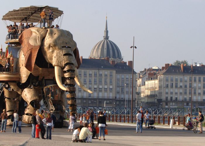 Un grand éléphant mécanique transportant des personnes sur son dos traverse une place publique, sous le regard de groupes de personnes qui prennent des photos. Des bâtiments européens classiques et une église à coupole sont visibles à l'arrière-plan.
