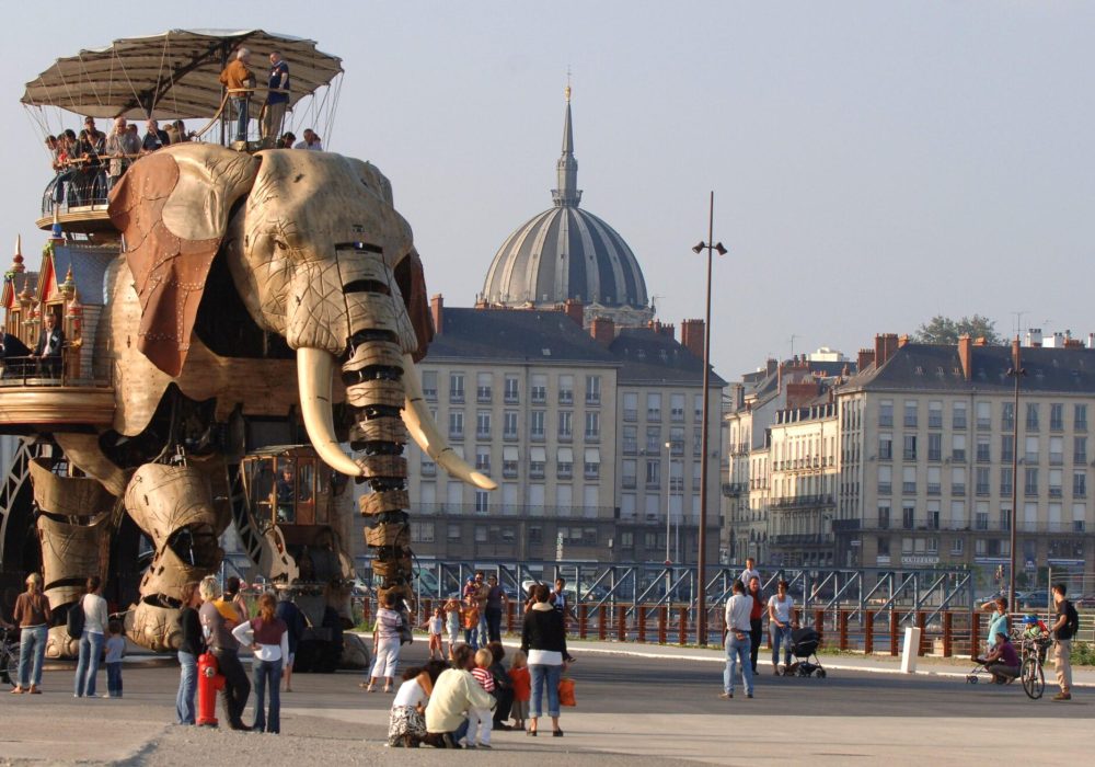 Un grand éléphant mécanique transportant des personnes sur son dos traverse une place publique, sous le regard de groupes de personnes qui prennent des photos. Des bâtiments européens classiques et une église à coupole sont visibles à l'arrière-plan.