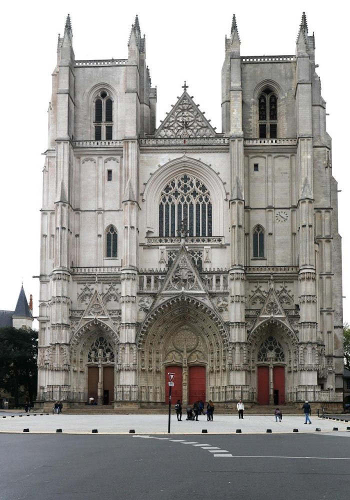 A large Gothic cathedral with two tall towers, intricate stone carvings, arched doorways and large stained glass windows. Several people stand near the entrance and the street is visible in the foreground.