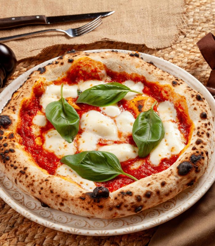 Une pizza Margherita de style napolitain avec de la mozzarella fondue, de la sauce tomate et des feuilles de basilic frais est posée sur une assiette à motifs à côté de moulins à poivre, de couverts et d'une serviette brune sur un tapis tissé.