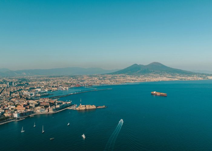 Aerial view of a coastal town with boats in the blue sea, a yellow ship and a distant volcano with two peaks under a clear blue sky.