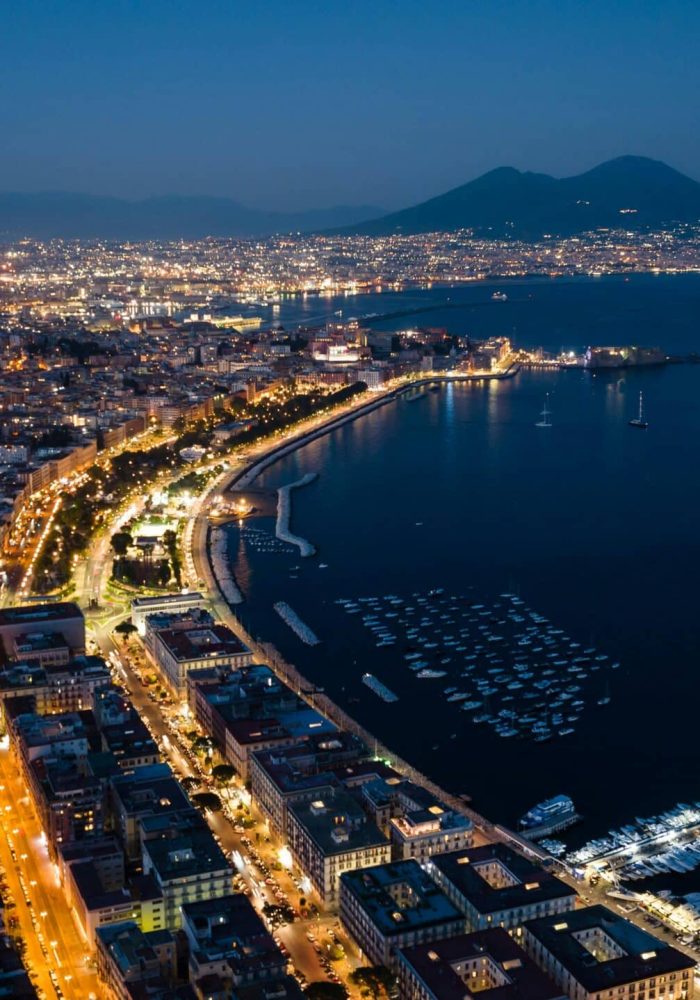 Aerial view of Naples, Italy, at dusk, with the lights of the city shining along the coast, boats anchored in the bay and Mount Vesuvius in the background, under a deep blue sky.