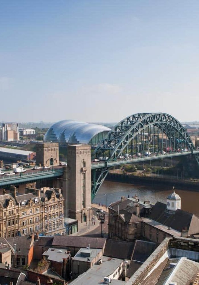 Panoramic view of Newcastle upon Tyne with Tyne Bridge spanning the river, historic buildings in the foreground and the modern Sage Gateshead concert hall in the background, under a clear blue sky.