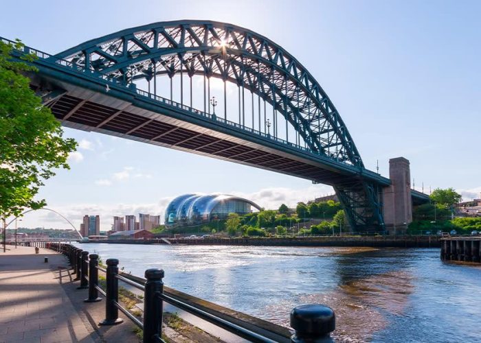 Un grand pont en arc enjambe une rivière par une journée ensoleillée, avec des bâtiments modernes et des arbres verts le long de la rive et un ciel bleu clair en arrière-plan.