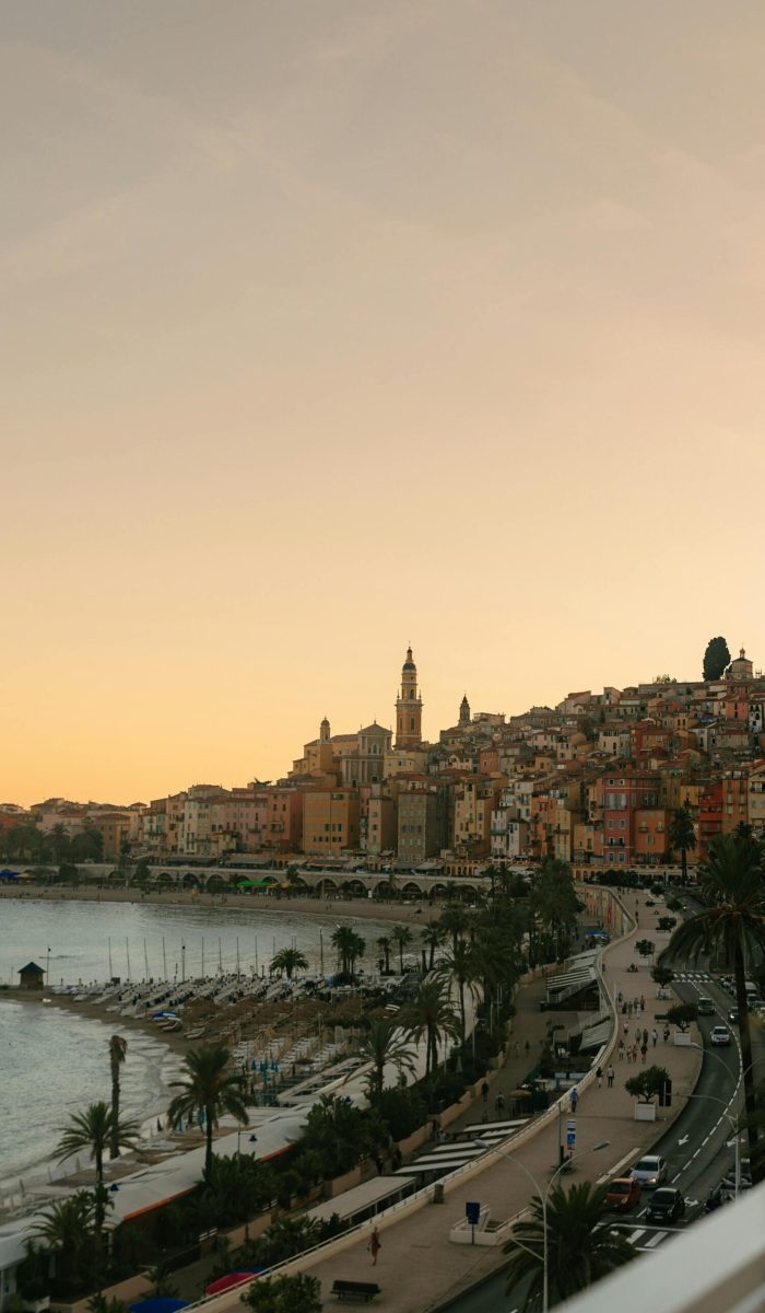 Une ville côtière au coucher du soleil avec des bâtiments aux couleurs pastel sur une colline, un clocher d'église, des palmiers le long de la plage et une route sinueuse le long du rivage. Le ciel s'illumine doucement de teintes chaudes.