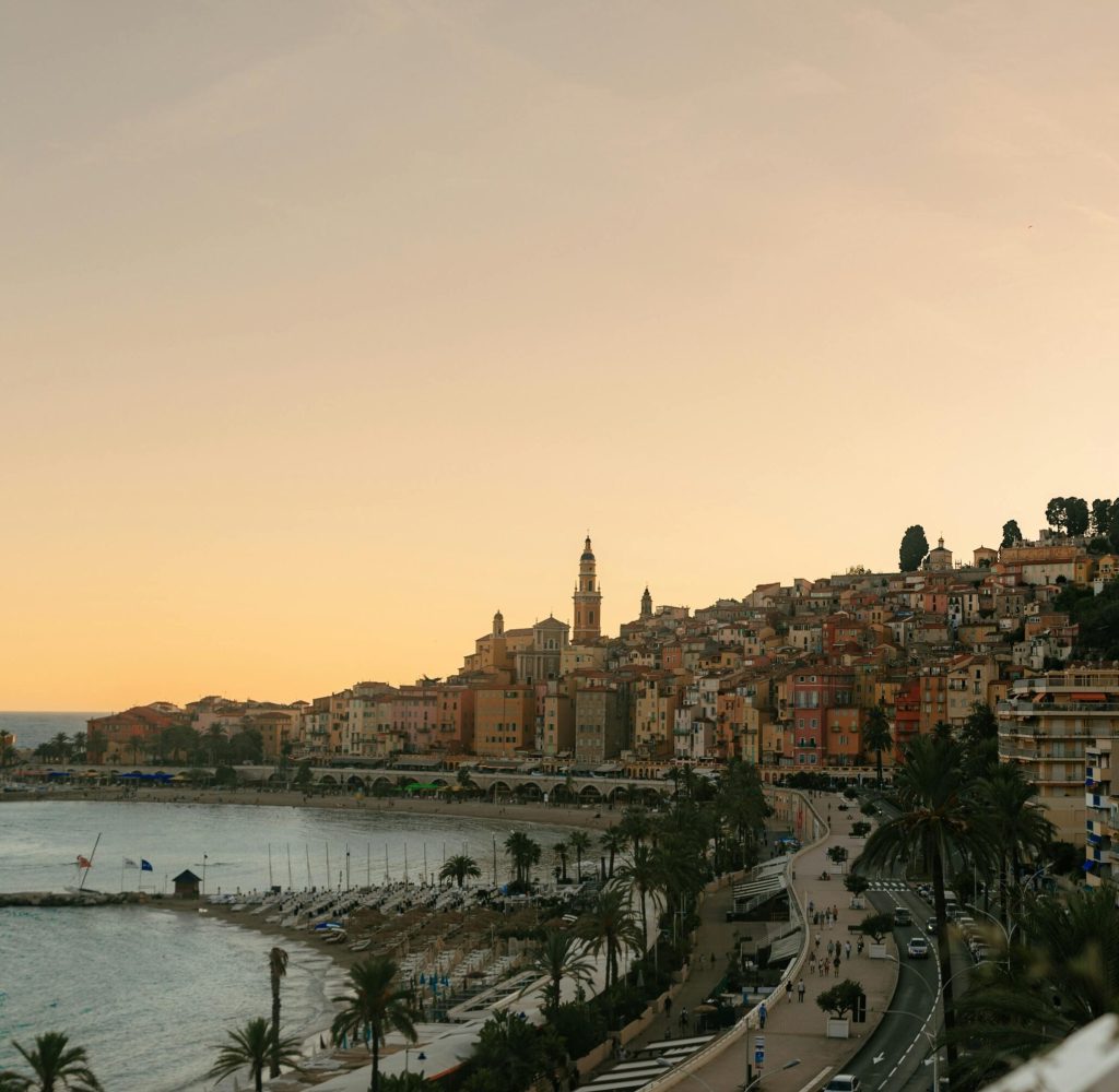 Une ville côtière au coucher du soleil avec des bâtiments aux couleurs pastel sur une colline, un clocher d'église, des palmiers le long de la plage et une route sinueuse le long du rivage. Le ciel s'illumine doucement de teintes chaudes.