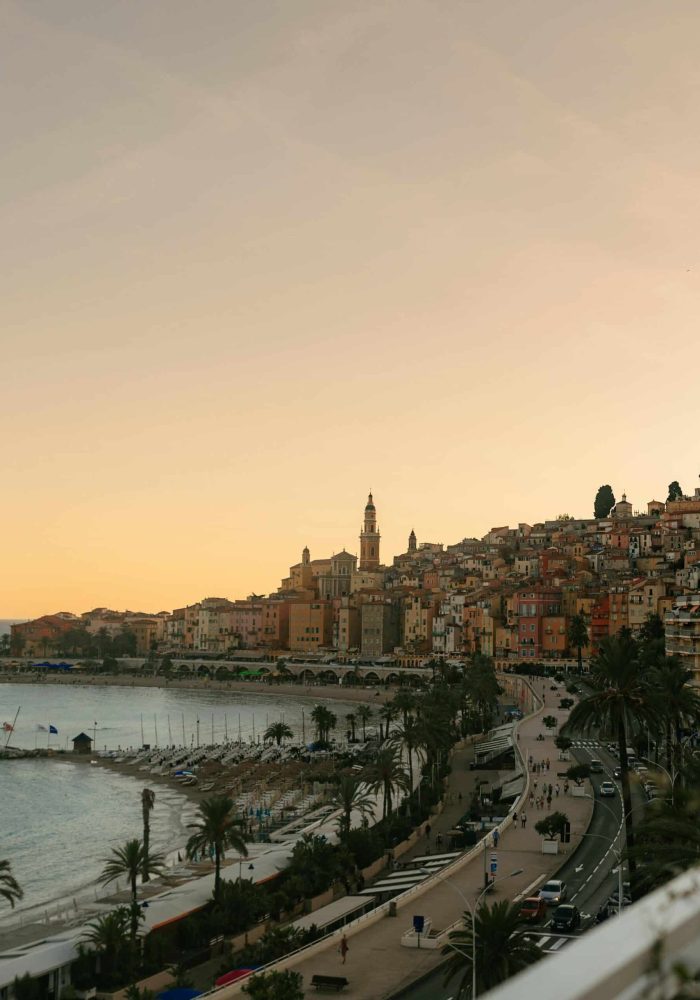 A coastal town at sunset with pastel-coloured buildings on a hill, a church steeple, palm trees along the beach and a winding road along the shore. The sky glows softly in warm hues.
