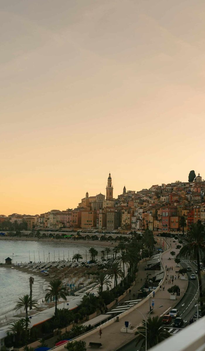 Une ville côtière au coucher du soleil avec des bâtiments aux couleurs pastel sur une colline, un clocher d'église, des palmiers le long de la plage et une route sinueuse le long du rivage. Le ciel s'illumine doucement de teintes chaudes.