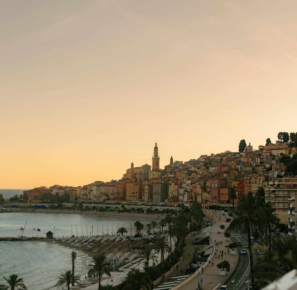 Une ville côtière au coucher du soleil avec des bâtiments aux couleurs pastel sur une colline, un clocher d'église, des palmiers le long de la plage et une route sinueuse le long du rivage. Le ciel s'illumine doucement de teintes chaudes.