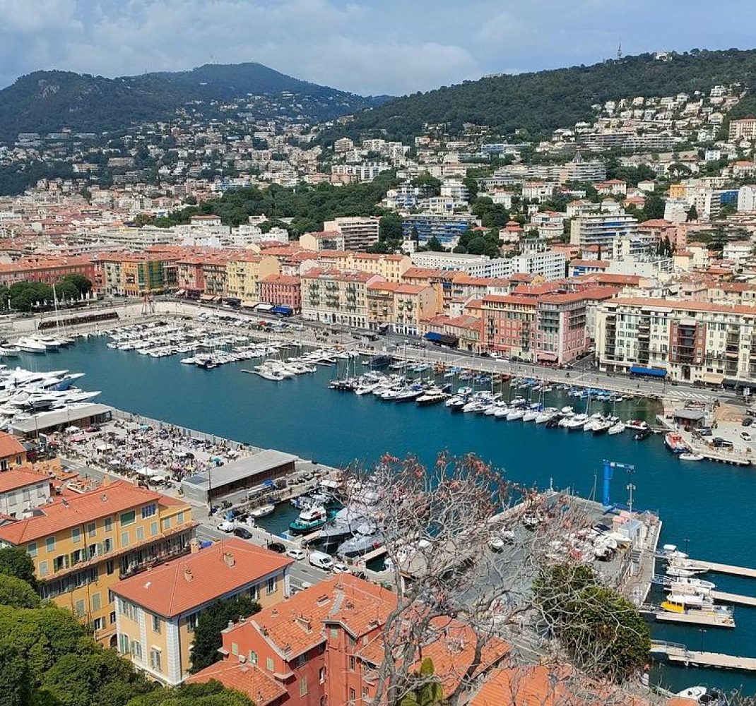 Vue aérienne d'un port de plaisance avec des yachts et des bateaux à quai, entouré de bâtiments colorés et adossé à des collines verdoyantes, dans une ville côtière méditerranéenne.