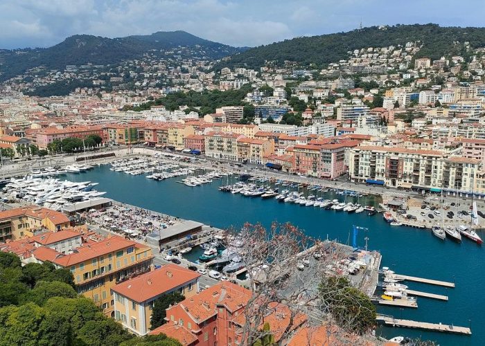 Aerial view of a marina with yachts and boats docked, surrounded by colourful buildings and backed by green hills, in a Mediterranean coastal town.