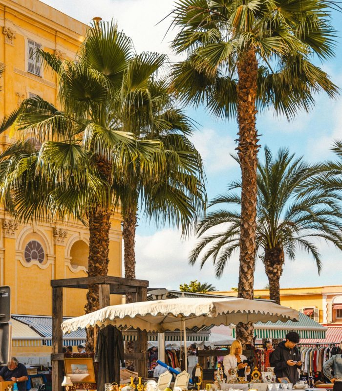 Un marché en plein air animé avec des tentes blanches, des tables exposant diverses marchandises et des gens qui se promènent. De grands palmiers et un bâtiment jaune aux fenêtres cintrées se trouvent à l'arrière-plan, sous un ciel partiellement nuageux.