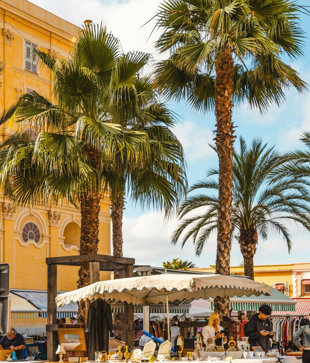 Un marché en plein air animé avec des tentes blanches, des tables exposant diverses marchandises et des gens qui se promènent. De grands palmiers et un bâtiment jaune aux fenêtres cintrées se trouvent à l'arrière-plan, sous un ciel partiellement nuageux.