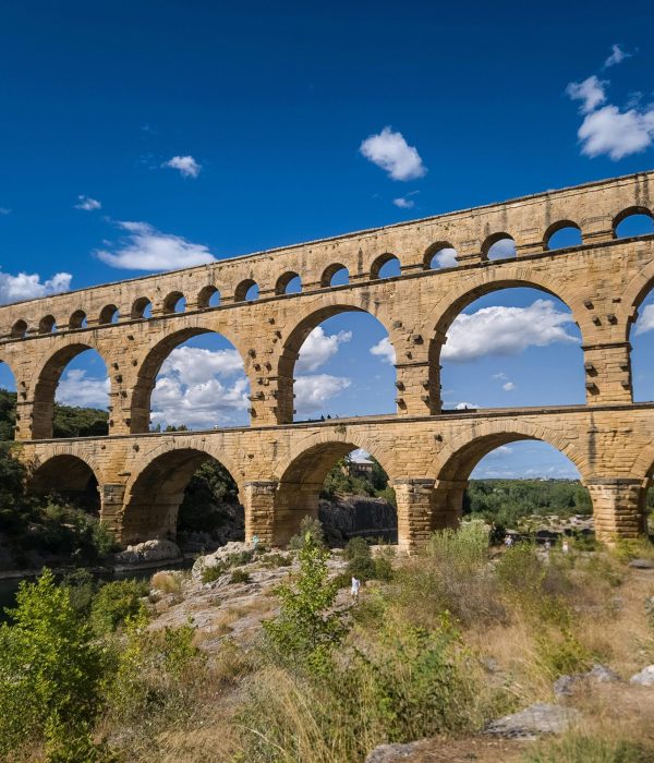Ancien aqueduc de pierre aux multiples arches enjambant une rivière, entouré de verdure et d'un ciel d'un bleu éclatant avec des nuages épars.