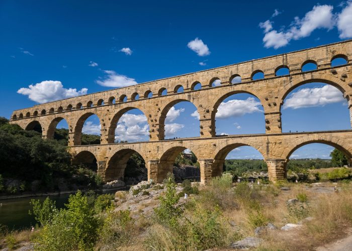 Ancien aqueduc de pierre aux multiples arches enjambant une rivière, entouré de verdure et d'un ciel d'un bleu éclatant avec des nuages épars.