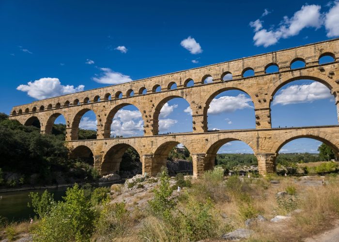 An ancient stone aqueduct with multiple arches spanning a river, surrounded by greenery and a brilliant blue sky with scattered clouds.