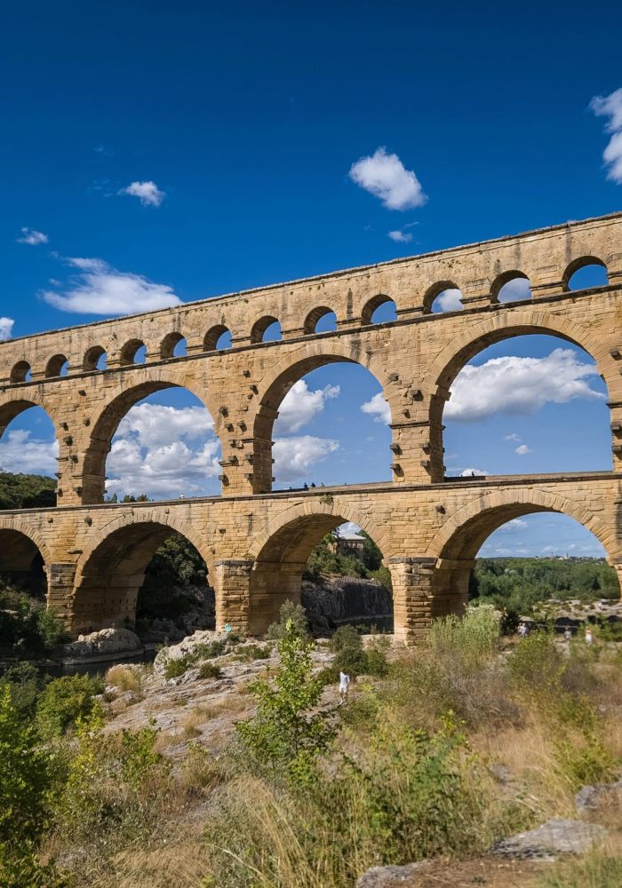 Ancien aqueduc de pierre aux multiples arches enjambant une rivière, entouré de verdure et d'un ciel d'un bleu éclatant avec des nuages épars.