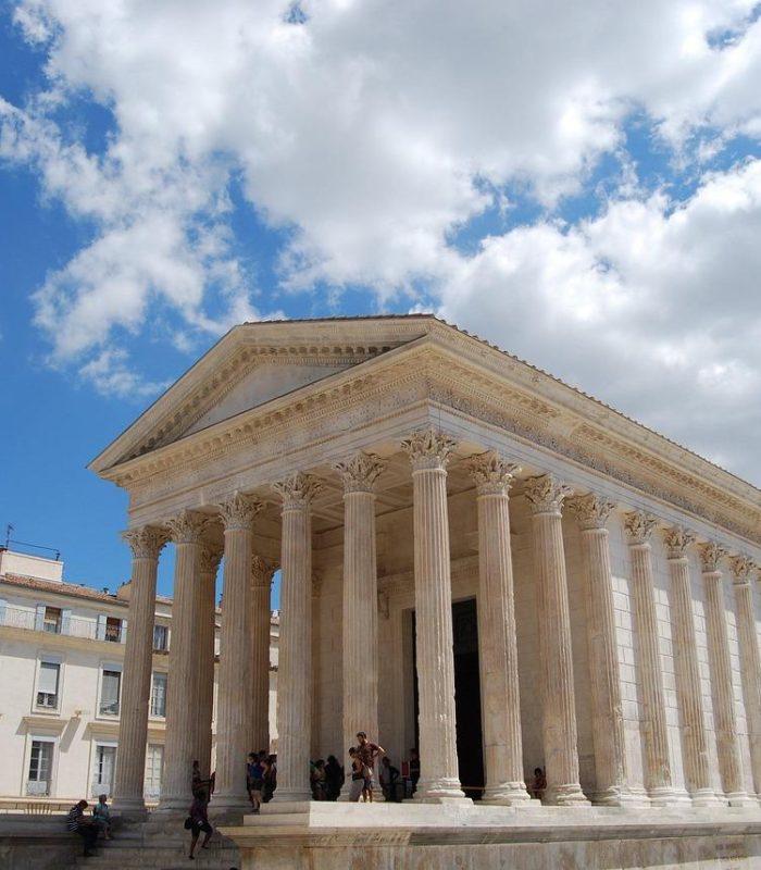 Temple romain antique avec de hautes colonnes et un fronton triangulaire, situé sur une place ensoleillée entourée de bâtiments modernes, sous un ciel bleu avec des nuages épars. Des personnes sont rassemblées sur les marches du temple.