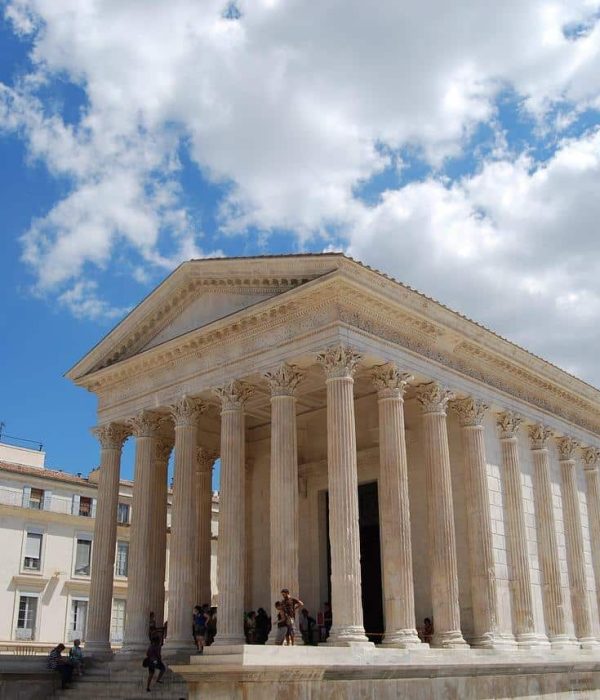 Temple romain antique avec de hautes colonnes et un fronton triangulaire, situé sur une place ensoleillée entourée de bâtiments modernes, sous un ciel bleu avec des nuages épars. Des personnes sont rassemblées sur les marches du temple.