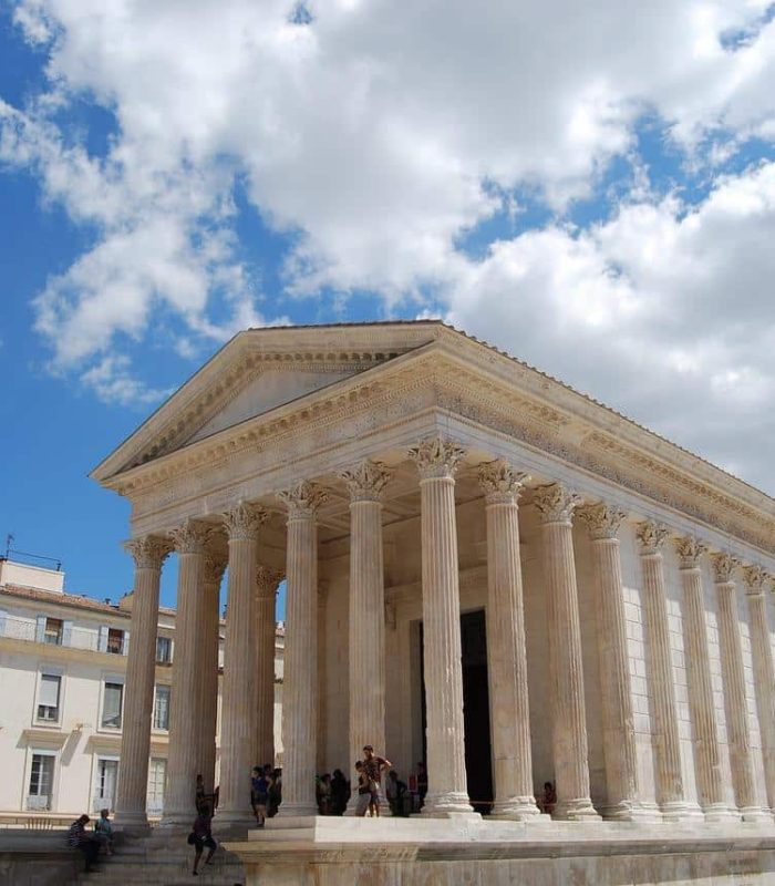 Ancient Roman temple with tall columns and a triangular pediment, situated in a sunny square surrounded by modern buildings, under a blue sky with scattered clouds. People are gathered on the steps of the temple.