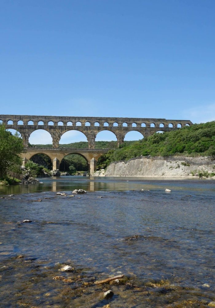 A large old stone aqueduct with multiple arches spans a river under a clear blue sky, with greenery and a stone building on the bank and a person walking nearby.