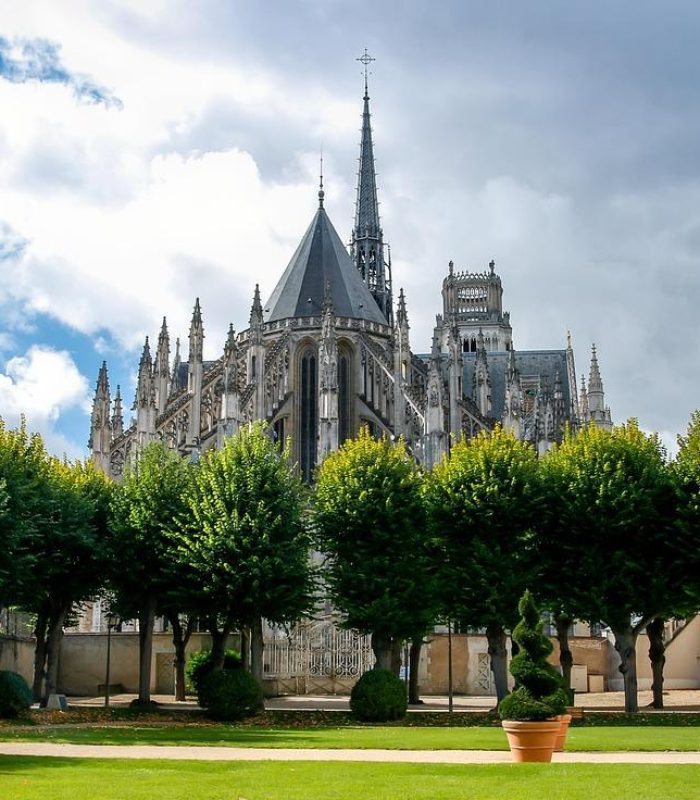 A Gothic cathedral with high spires rises behind a row of green trees in a manicured garden, under a partly cloudy sky.