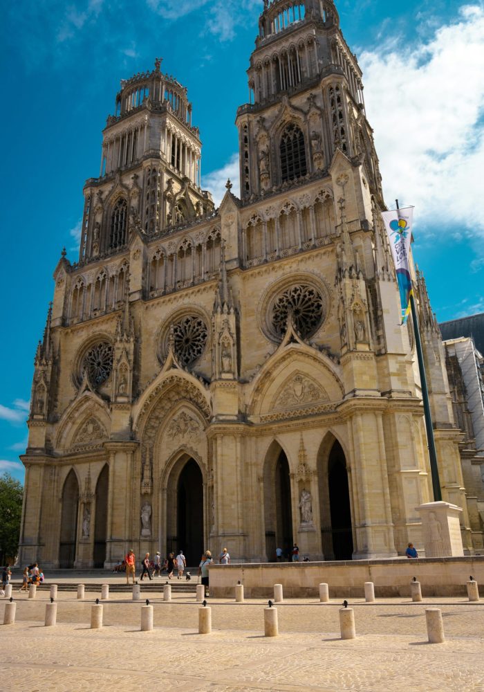 Une grande cathédrale gothique avec deux hautes tours, des arcs ornés et des vitraux circulaires, sur fond de ciel bleu, avec des gens rassemblés autour de l'entrée.