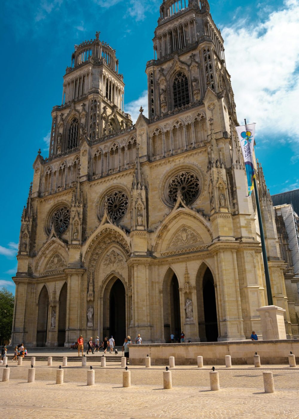 Une grande cathédrale gothique avec deux hautes tours, des arcs ornés et des vitraux circulaires, sur fond de ciel bleu, avec des gens rassemblés autour de l'entrée.
