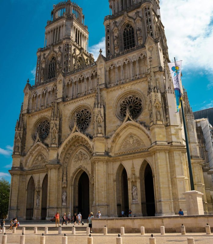 A large Gothic cathedral with two tall towers, ornate arches and circular stained glass windows, against a backdrop of blue sky, with people gathered around the entrance.