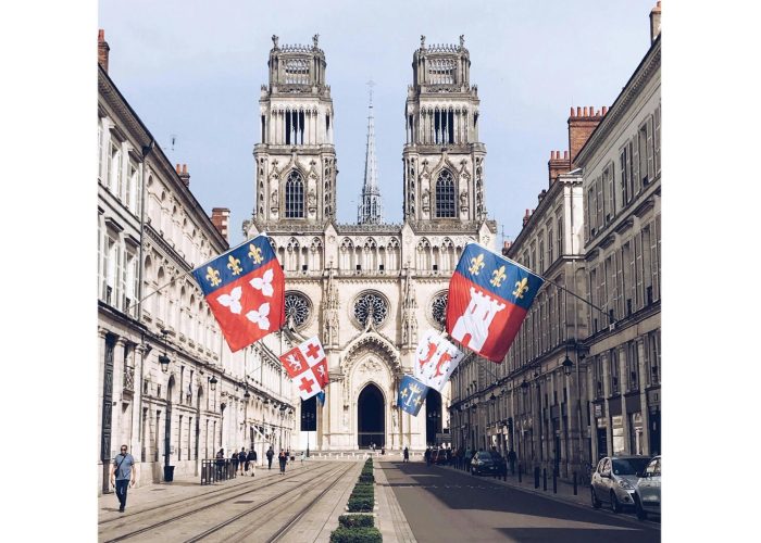 Une large rue bordée de bâtiments élégants mène à une grande cathédrale gothique à deux tours. Des drapeaux colorés sont suspendus au-dessus de la rue, et quelques personnes se promènent sur les trottoirs par temps clair.