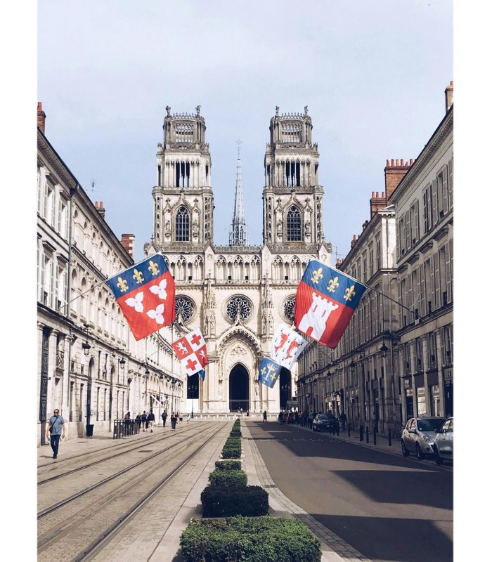 Une large rue bordée de bâtiments élégants mène à une grande cathédrale gothique à deux tours. Des drapeaux colorés sont suspendus au-dessus de la rue, et quelques personnes se promènent sur les trottoirs par temps clair.