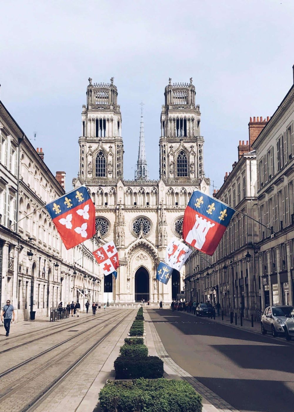 Une large rue bordée de bâtiments élégants mène à une grande cathédrale gothique à deux tours. Des drapeaux colorés sont suspendus au-dessus de la rue, et quelques personnes se promènent sur les trottoirs par temps clair.