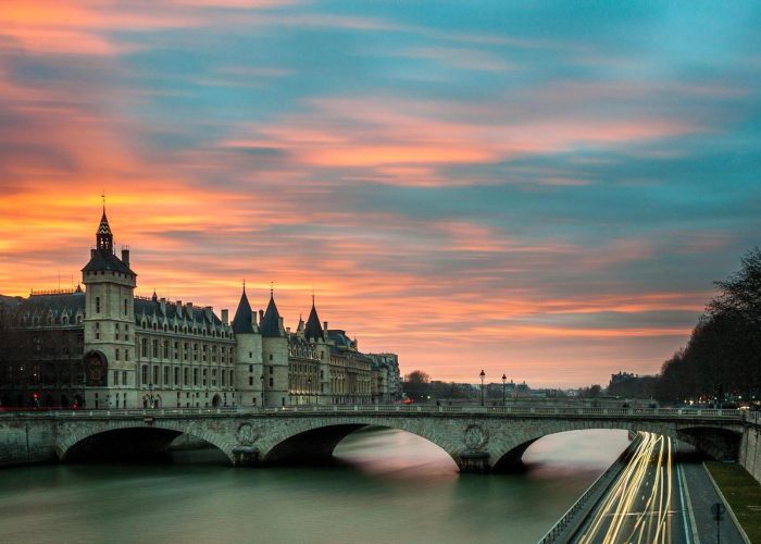 Panoramic view of the Conciergerie and the Pont au Change on the Seine in Paris at sunset, with coloured clouds in the sky and streaks of light from vehicles passing along the riverside road.
