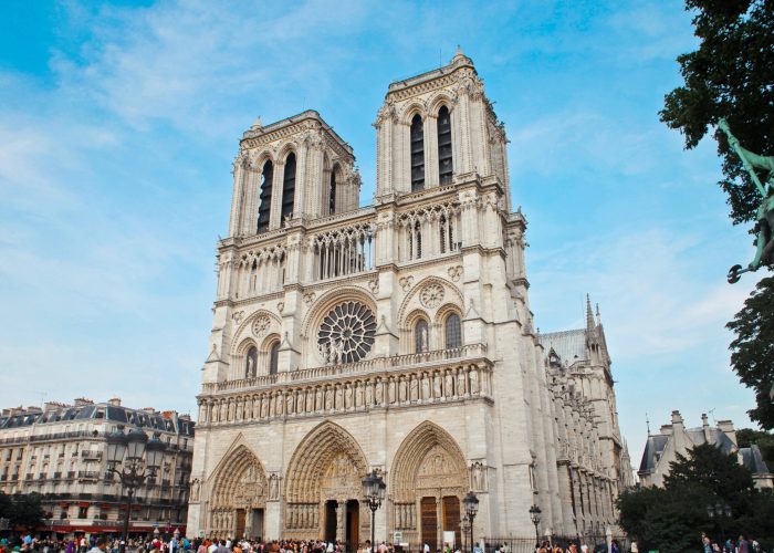 La cathédrale Notre-Dame de Paris, avec ses tours jumelles emblématiques, sa façade ornée et sa grande rosace, se détache sur un ciel bleu alors que des foules se rassemblent à son entrée.