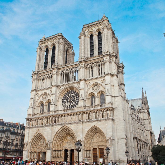 La cathédrale Notre-Dame de Paris, avec ses tours jumelles emblématiques, sa façade ornée et sa grande rosace, se détache sur un ciel bleu alors que des foules se rassemblent à son entrée.
