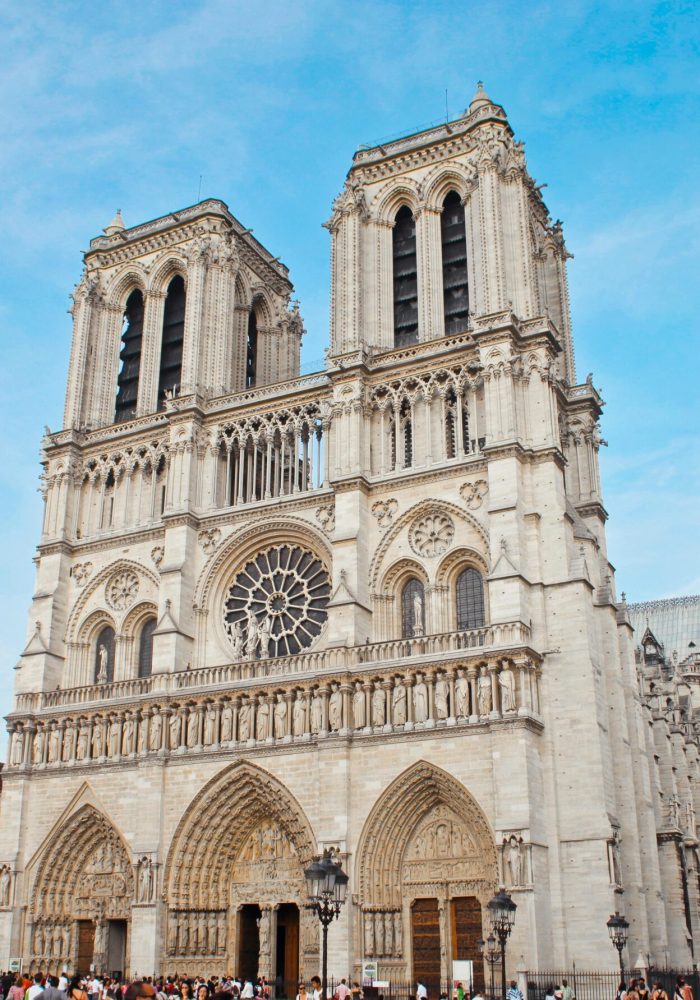 Notre-Dame de Paris cathedral, with its iconic twin towers, ornate façade and large rose window, stands out against a blue sky as crowds gather at its entrance.