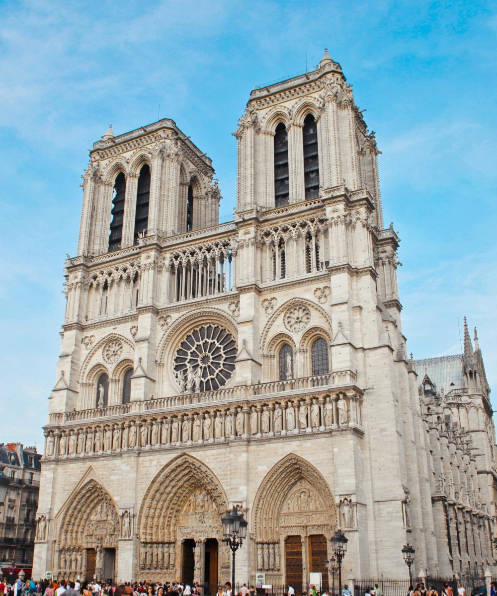 La cathédrale Notre-Dame de Paris, avec ses tours jumelles emblématiques, sa façade ornée et sa grande rosace, se détache sur un ciel bleu alors que des foules se rassemblent à son entrée.