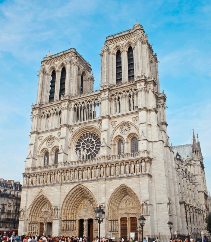 Notre-Dame de Paris cathedral, with its iconic twin towers, ornate façade and large rose window, stands out against a blue sky as crowds gather at its entrance.